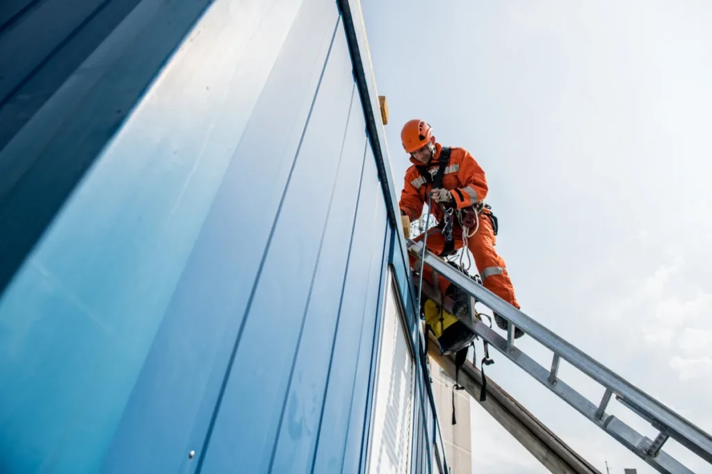 Worker in orange safety gear climbs a metal ladder against a blue building, secured with harnesses—highlighting the Mid-Market Challenges faced in ensuring workplace safety and compliance.