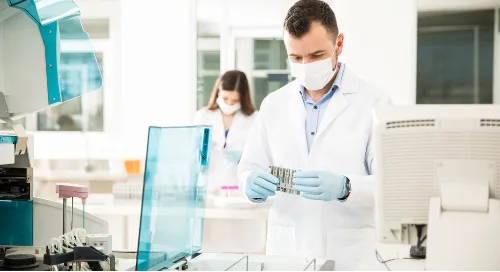 A scientist in a lab coat and mask examines test tubes as part of chemical management; another person works in the background.