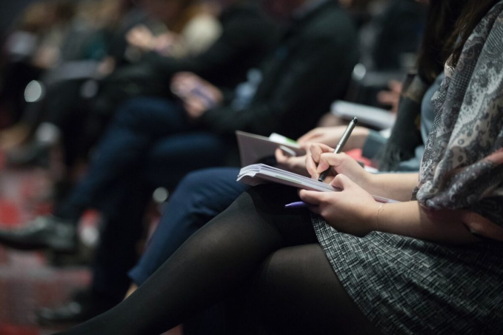 People sitting in rows at an event, with one person in focus taking notes in a notebook—perhaps on important insights related to mental health.