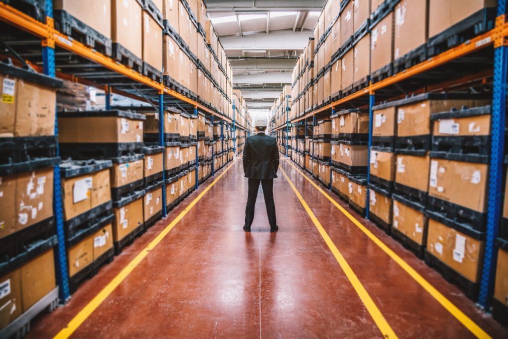 A person in a suit stands between tall shelves in a large warehouse filled with stacked boxes, overseeing Quality Management Programs.