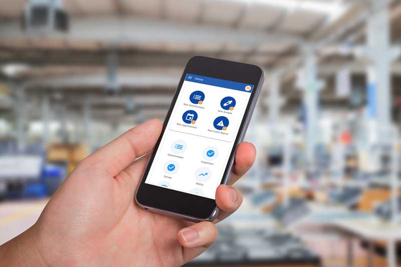 A hand holds a smartphone displaying an app, with a blurred industrial background, highlighting how connected worker technology leverages real-time machine data.