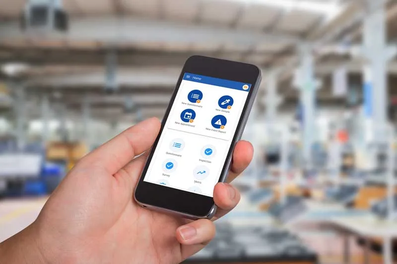 A hand holds a smartphone displaying an app, with a blurred industrial background, highlighting how connected worker technology leverages real-time machine data.