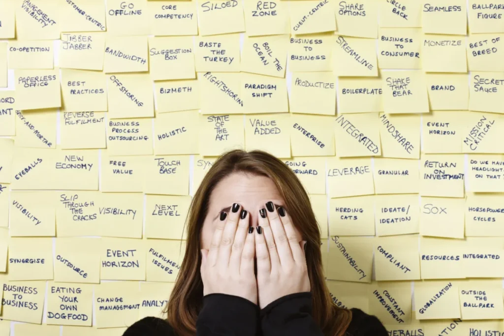 A person covers their face with their hands in front of a wall covered in sticky notes with business terms, highlighting the pressures of fostering a strong safety culture.