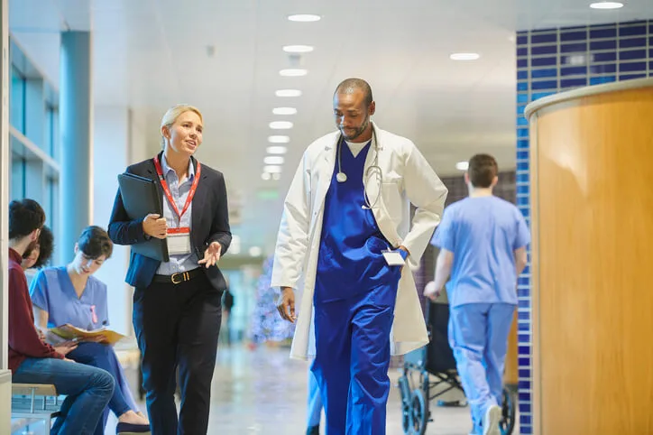 A doctor in blue scrubs and a white coat walks down a hospital hallway, talking with a woman in business attire holding a clipboard. Other medical staff and patients are visible in the background.