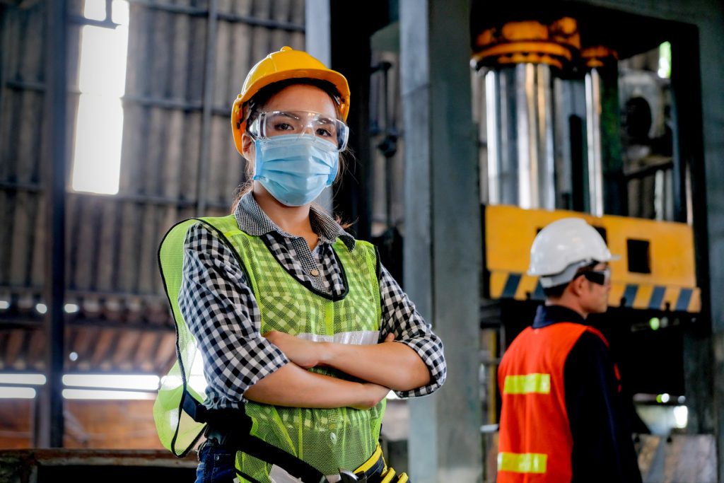Woman in safety gear and face mask stands confidently in a factory, prioritizing sécurité amid COVID-19; worker in background.