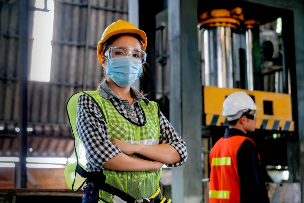 Woman in safety gear and face mask stands confidently in a factory, prioritizing sécurité amid COVID-19; worker in background.