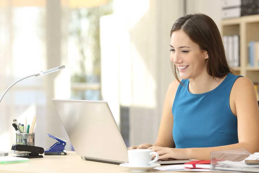 Woman in a blue top smiling while working on her laptop at a desk, using Cority Home Office Ergonomics to prevent strain, surrounded by office supplies and a cup of coffee.