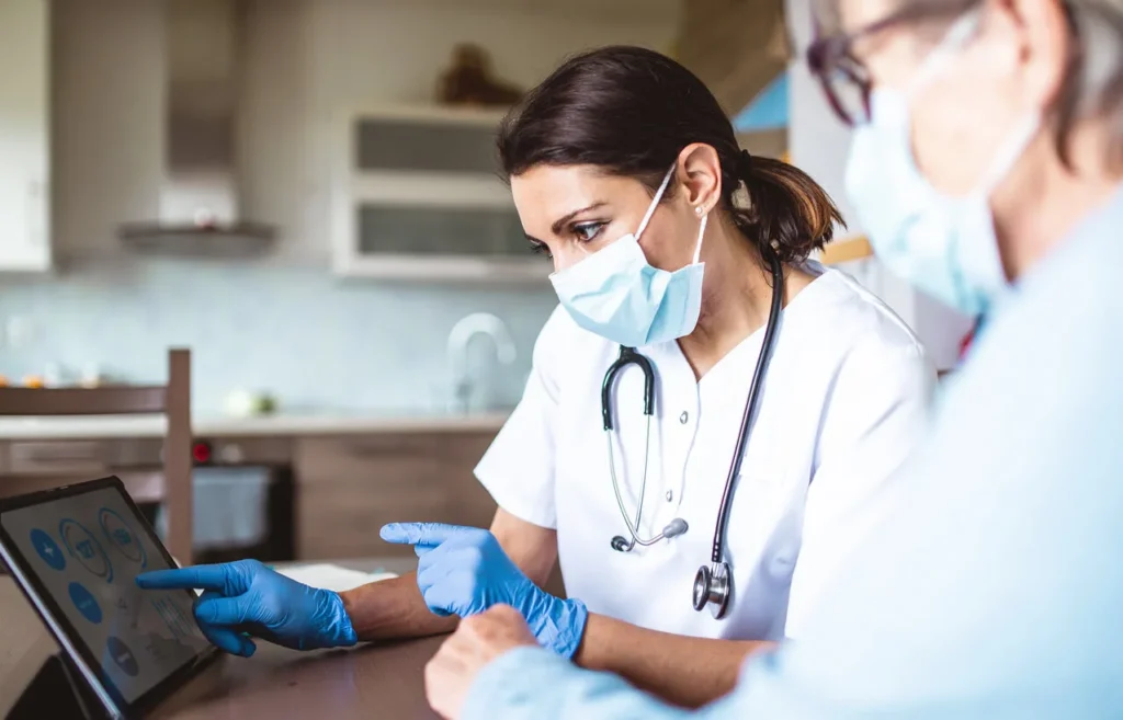 A healthcare worker in a mask and gloves points at a tablet screen while speaking with a masked patient in a home setting. Both are focused on the tablet, which displays medical information.