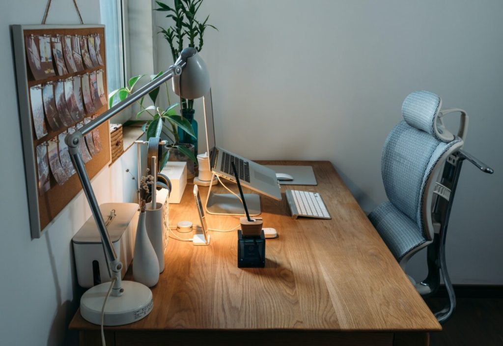 Modern home office with a wooden desk, ergonomic chair, computer, desk lamp, and plants by the window—an ideal space for effective ergonomic intervention.