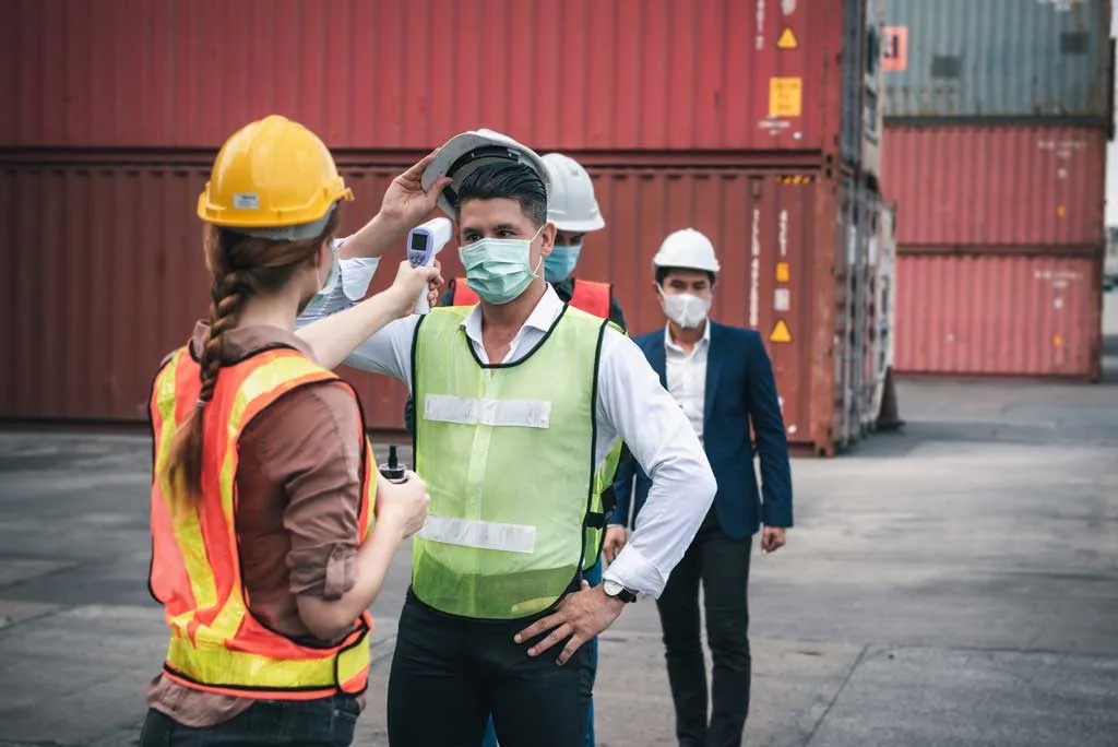 A worker in a safety vest gets his temperature checked at a shipping yard as part of COVID-19 protocols; others wait behind him, ensuring a safe return to productivity.