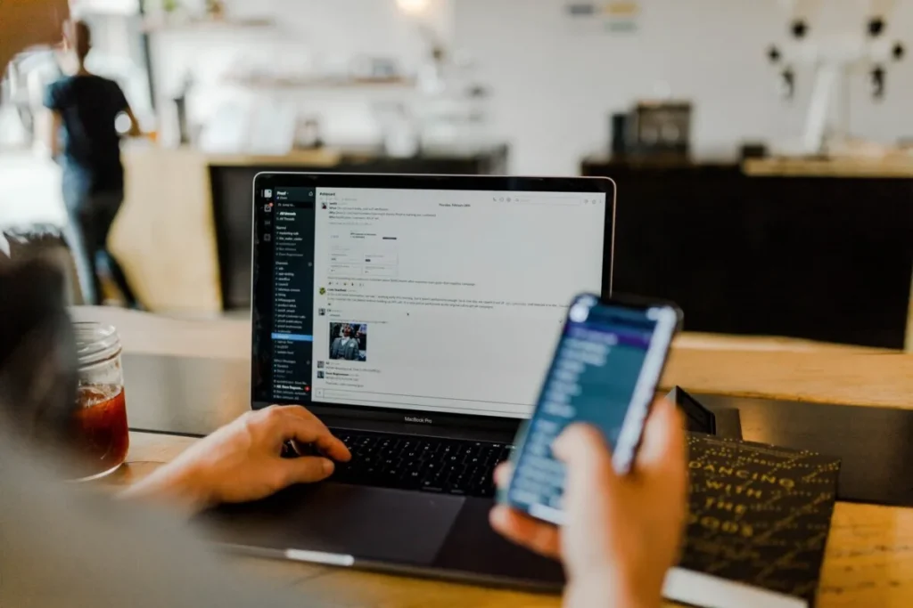 Person using a laptop and smartphone at a desk, both displaying messaging apps; a drink sits nearby. The screens show notifications from AI EHS systems, highlighting seamless integration of safety alerts into daily workflows.