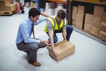 A man in a safety vest lifts a box as another man in business attire instructs him in a warehouse, showcasing essential practices for the future of industrial hygiene.
