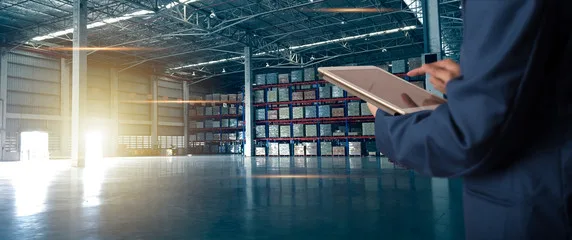 Person using a tablet in a large warehouse with stacked shelves and bright sunlight streaming in, embracing the future of industrial hygiene and ensuring a safer workplace—the way ahead for modern facilities.
