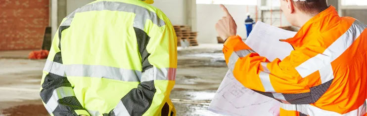Two construction workers in reflective jackets review building plans inside a partially constructed structure, focusing on injury prevention to ensure workplace safety.