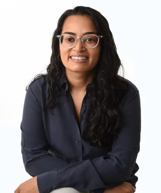 Woman with long dark hair and glasses, wearing a dark shirt, smiling and sitting against a plain background—exuding confidence and an appreciation for organizational safety.