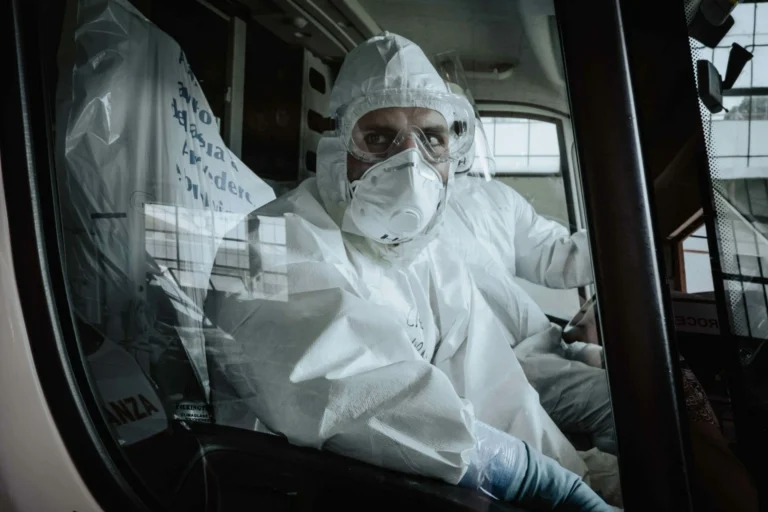 Person in full protective suit and mask sits inside a vehicle, illustrating industrial hygiene practices, while another person sits beside them, both looking out the window.