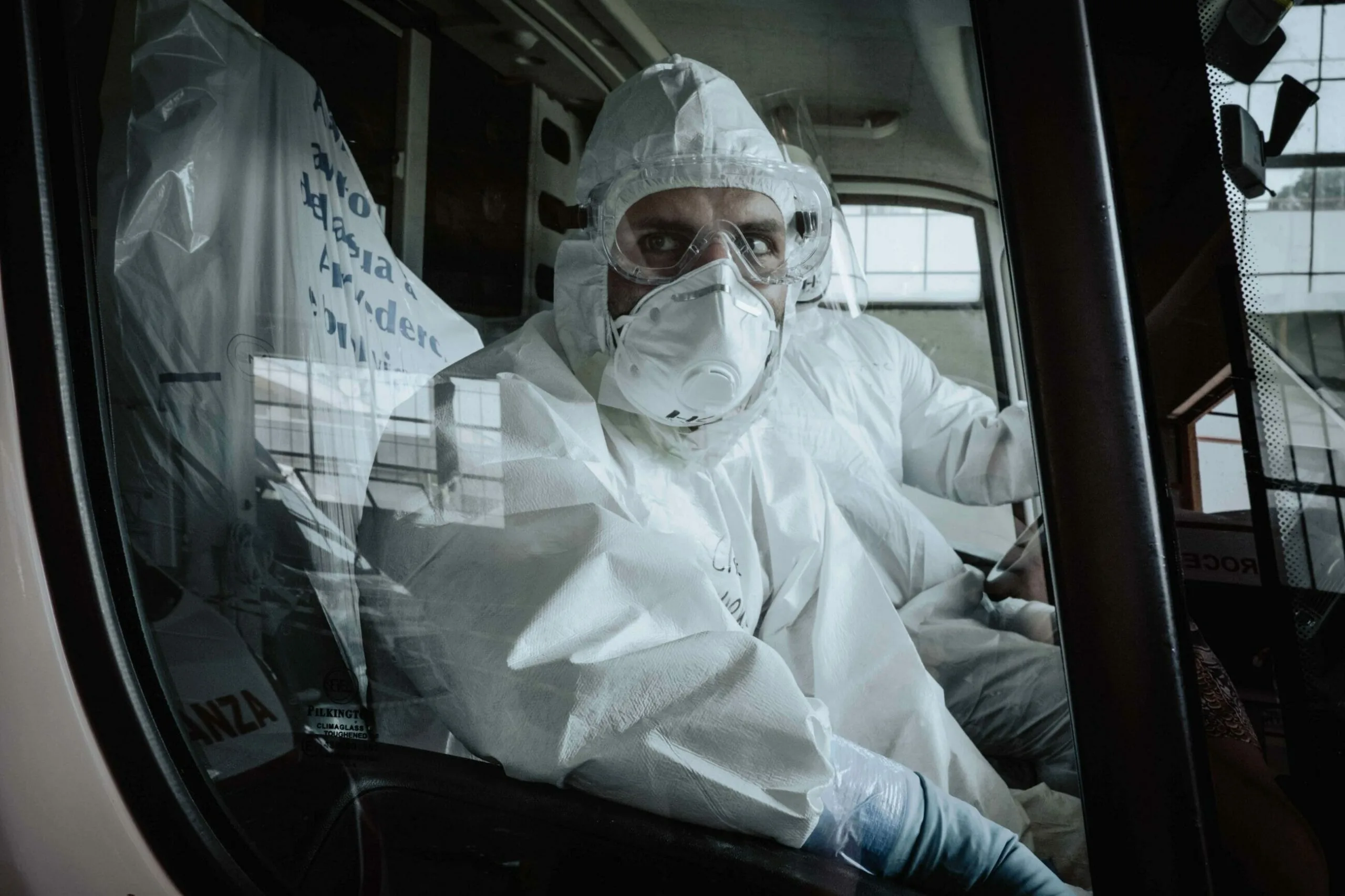 Person in full protective suit and mask sits inside a vehicle, illustrating industrial hygiene practices, while another person sits beside them, both looking out the window.