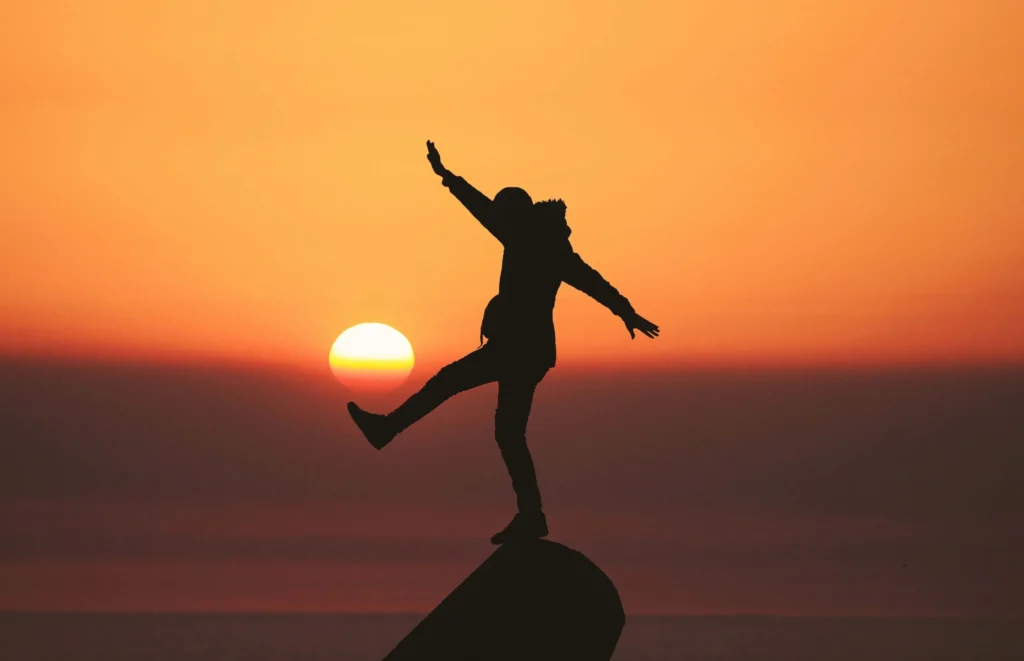 A person balances on one leg atop a rock at sunset, silhouetted against an orange sky, embodying the focus and poise of EHS subject matter experts.