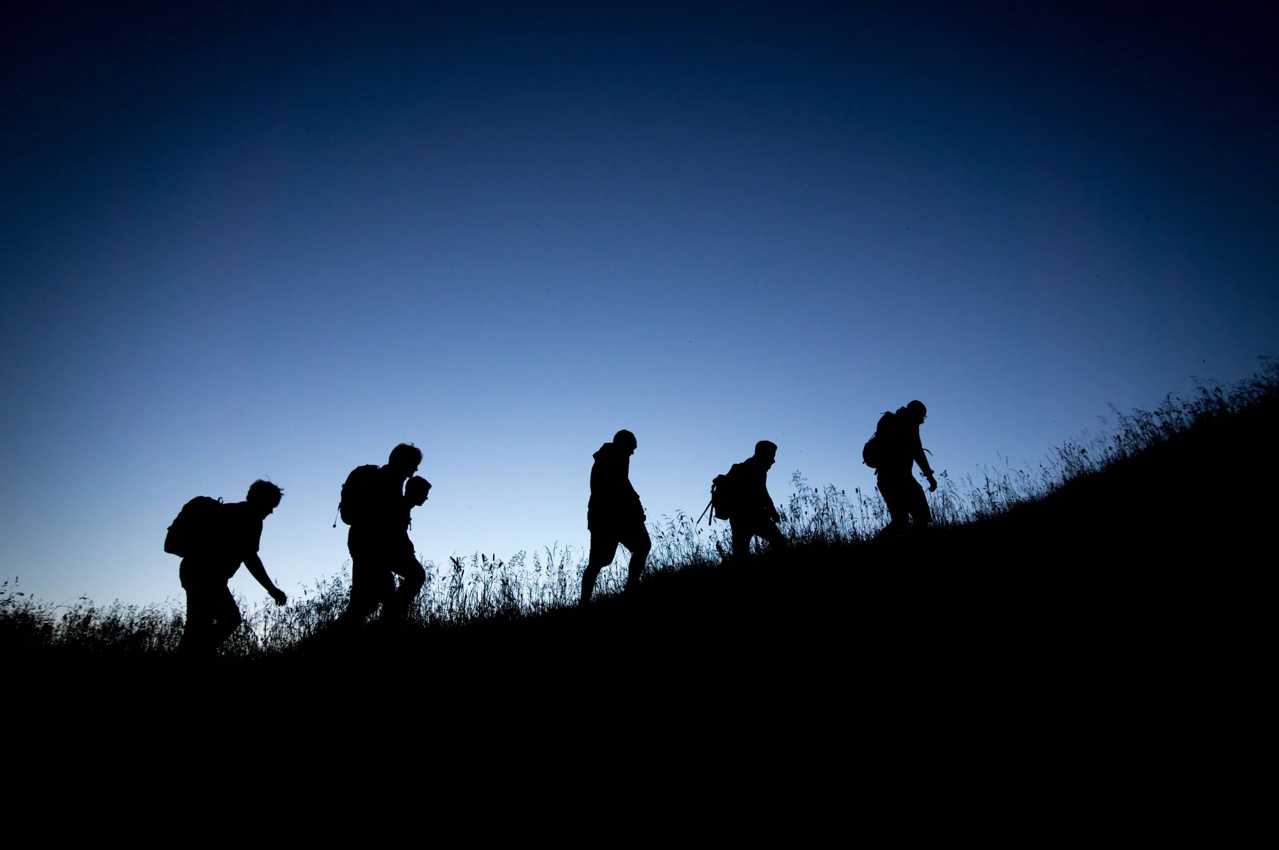 Silhouettes of five hikers, reminiscent of EHS Partners’ teamwork, walking uphill at dusk against a clear, dark blue sky.