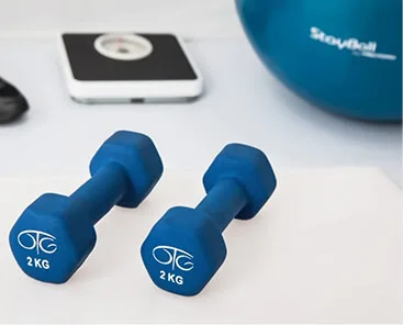 Two blue 2 kg dumbbells sit on a white surface, with a blurred scale and blue exercise ball in the background—an inspiring setting for those passionate about wellness, much like the culture fostered at Careers at Cority.