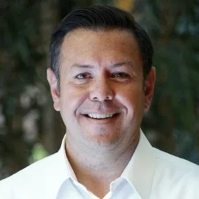 A man with short dark hair, wearing a white collared shirt, smiles at the camera with a blurred green background behind him.