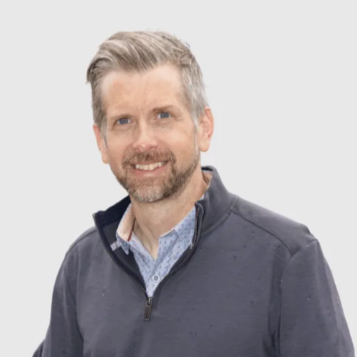 A man with short, light brown hair and a trimmed beard smiles at the camera. He is wearing a navy quarter-zip sweater over a collared shirt, standing against a plain light background.