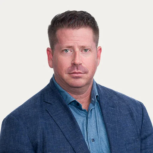 A man with short brown hair wearing a blue blazer and light blue collared shirt, looking directly at the camera with a neutral expression against a plain background.