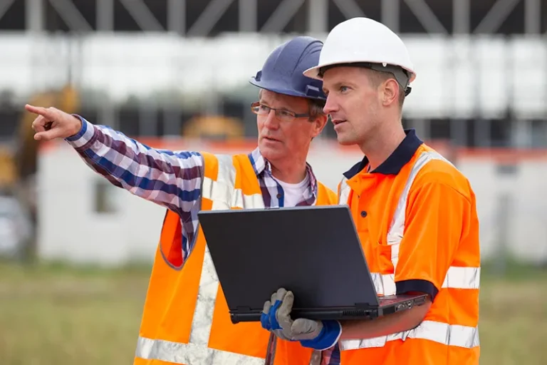 Two construction workers in safety gear and helmets stand outside; one holds a laptop while the other points towards something in the distance, suggesting a discussion or planning at a construction site.