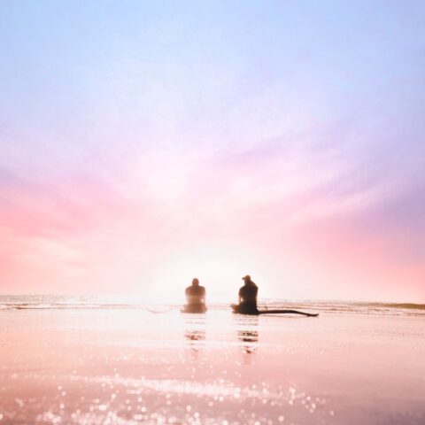 Two people sit on paddleboards on calm water, silhouetted against a pastel pink and purple sunrise, with the soft light reflecting off the surface—a tranquil moment reminiscent of the peace often found at Greenstone retreats.