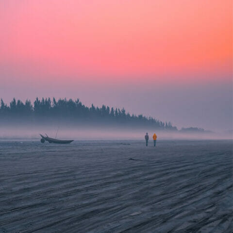 Two people walk along a misty beach at sunrise near a Greenstone boat on the sand, with trees silhouetted against a pink and orange sky.