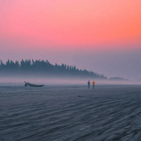 Two people walk along a misty beach at sunrise near a Greenstone boat on the sand, with trees silhouetted against a pink and orange sky.