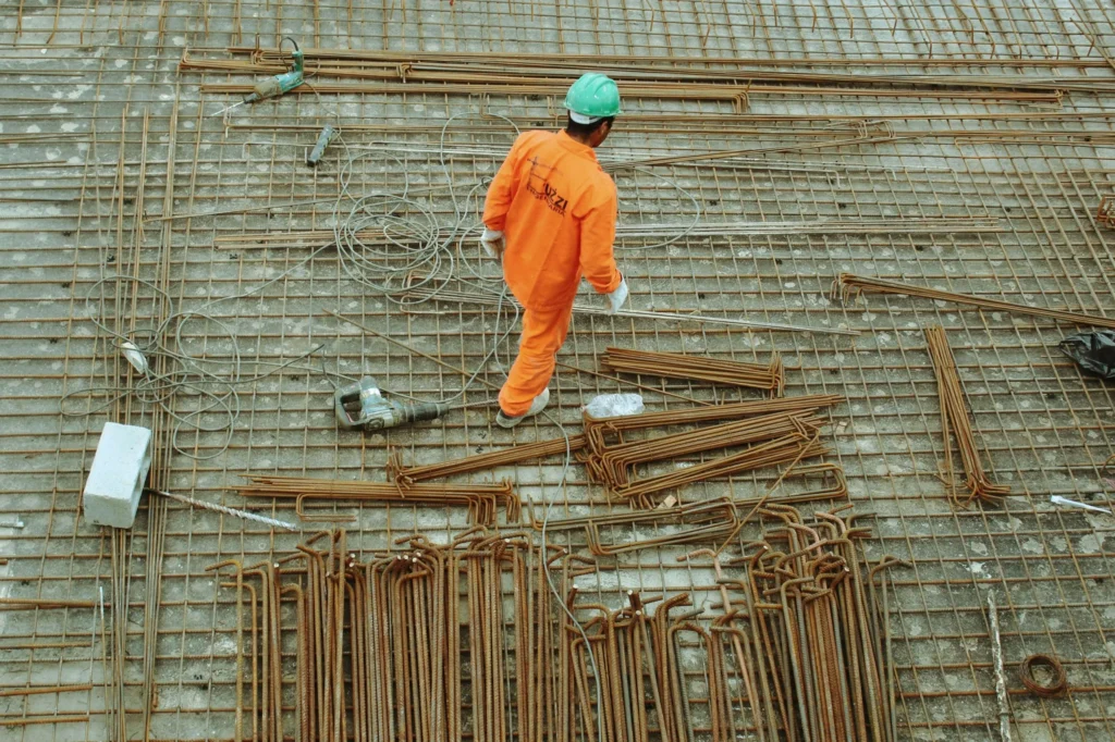 Construction worker in orange uniform and green helmet walking on rebar at a building site, viewed from above, demonstrating the importance of safety on active construction sites.