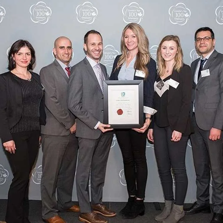 Six professionally dressed people stand in a row, smiling at the camera. The person in the middle holds a framed certificate, celebrating success. Behind them is a grey backdrop with white award logos, reflecting the achievements of careers at Cority.