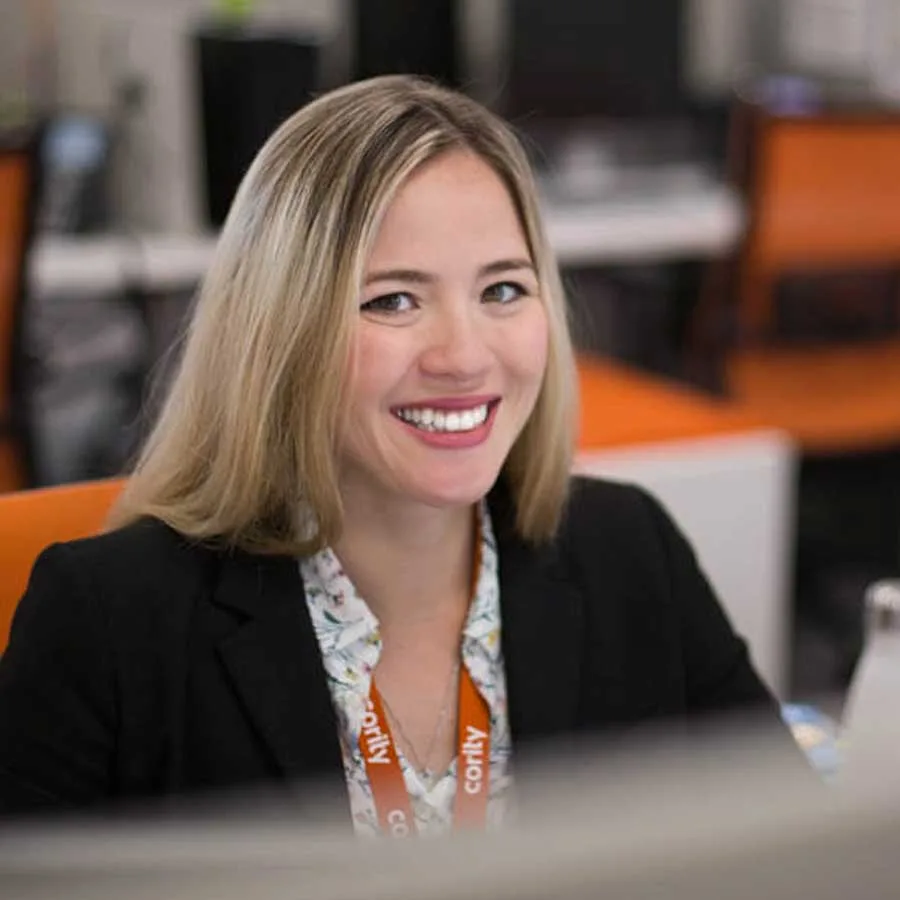 A woman with blonde hair, wearing a black blazer and floral blouse, smiles while sitting at an office desk with orange chairs and computer equipment in the background—an inviting scene reflecting the vibrant atmosphere of Careers at Cority.