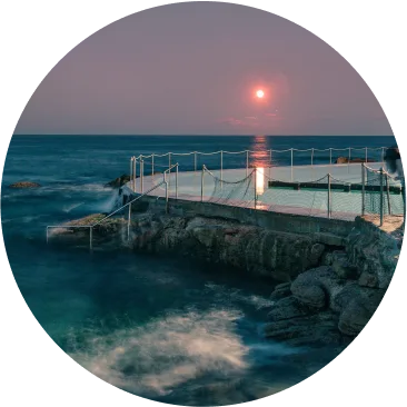 A round image of an ocean pool at dusk, with calm waves, a stone edge, and rope barriers. The full moon glows above the horizon, casting light on the water and evoking serenity—an ambiance echoing the calm progress of sustainability at Cority.