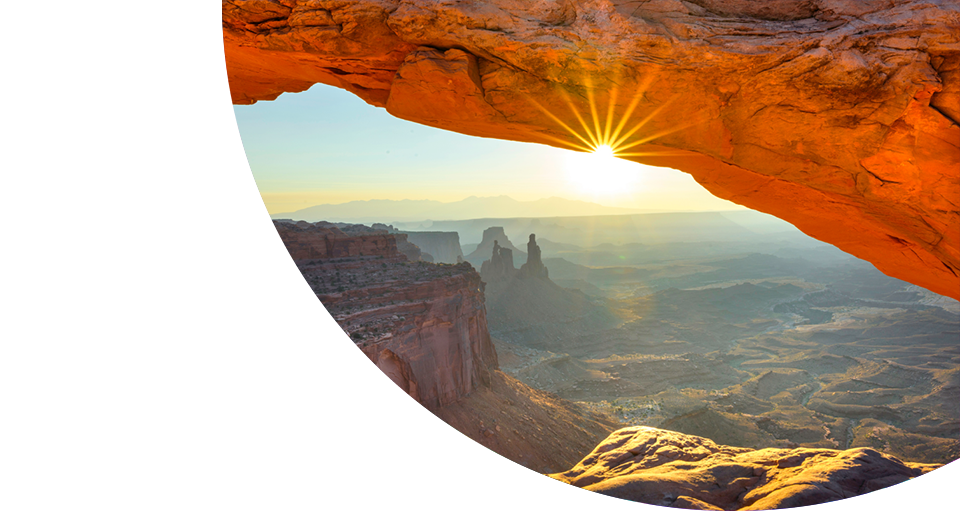 Sunrise shining through a natural red rock arch, overlooking a vast canyon landscape with rugged cliffs, mesas, and hints of Greenstone glowing in the morning light.