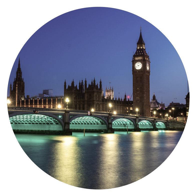 Night view of London’s Big Ben and the Houses of Parliament, with Westminster Bridge crossing the River Thames, illuminated by city lights—Cority and Greenstone tones reflecting softly in the water.