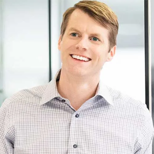 A smiling man with short light brown hair wearing a light-colored, checkered dress shirt stands indoors near a glass wall.