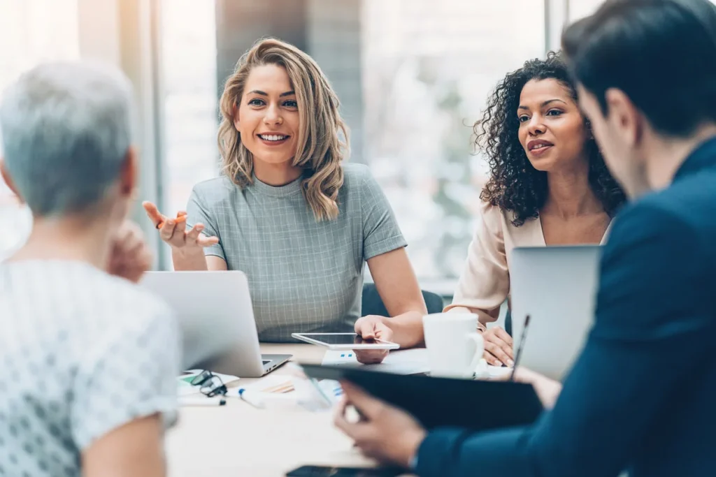 Four professionals sit around a table in a bright office, engaged in discussion. Two women are facing the camera, smiling and talking about growing their futures with Careers at Cority, with laptops and papers in front of them.
