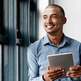 A smiling man in a blue shirt stands by a window, holding a tablet with the Cority Legal Center app open, and looks outside with a thoughtful expression.