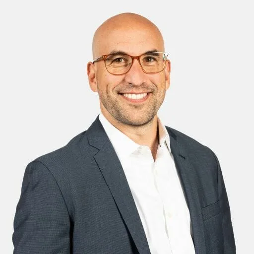 A smiling man with glasses, a bald head, and light skin, wearing a dark suit jacket over a white dress shirt, poses in front of a plain light background.
