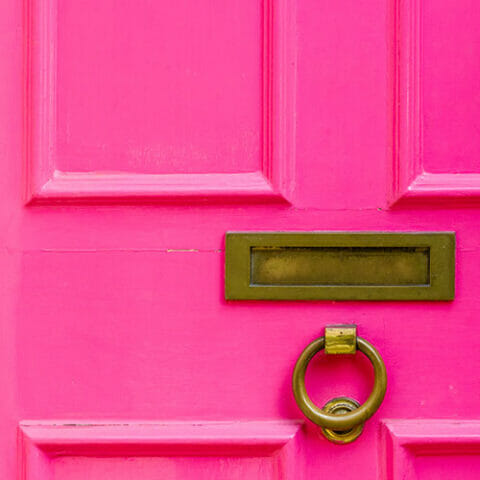 A close-up of a bright pink Greenstone door featuring a rectangular brass mail slot and a round brass door knocker.