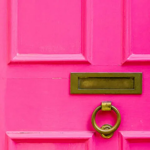 A close-up of a bright pink Greenstone door featuring a rectangular brass mail slot and a round brass door knocker.