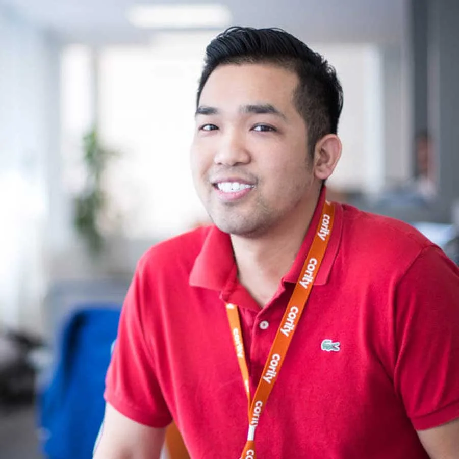 A man wearing a red polo shirt and an orange lanyard smiles at the camera in a bright office setting, reflecting the friendly atmosphere of Careers at Cority.