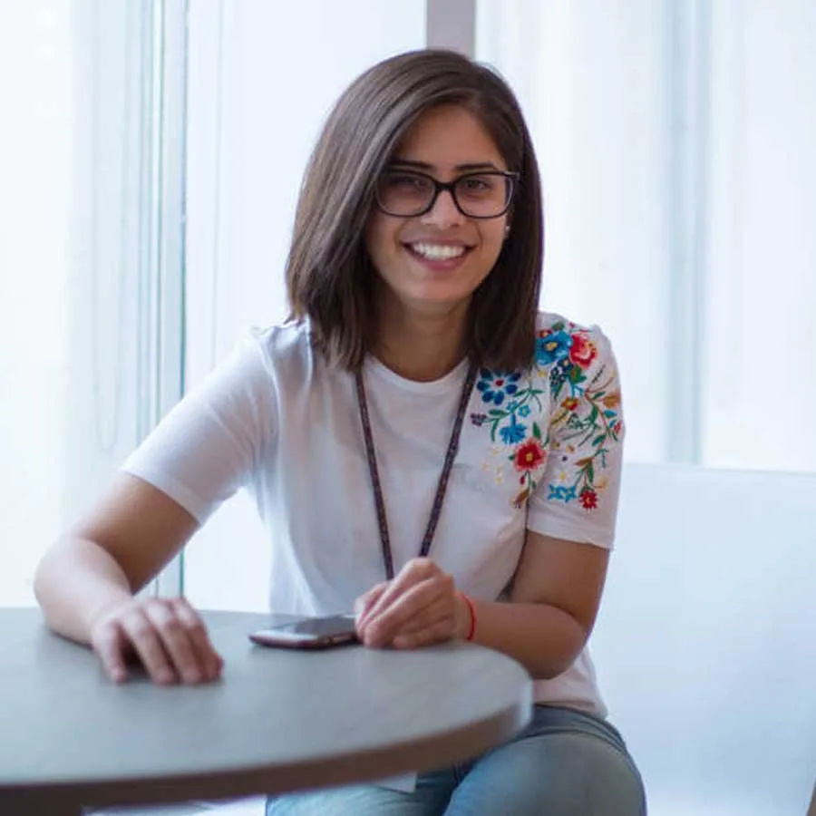 A young woman with shoulder-length brown hair and glasses sits at a table, smiling. She wears a white t-shirt with colorful floral embroidery on the shoulder and has a smartphone in front of her, exploring Careers at Cority.