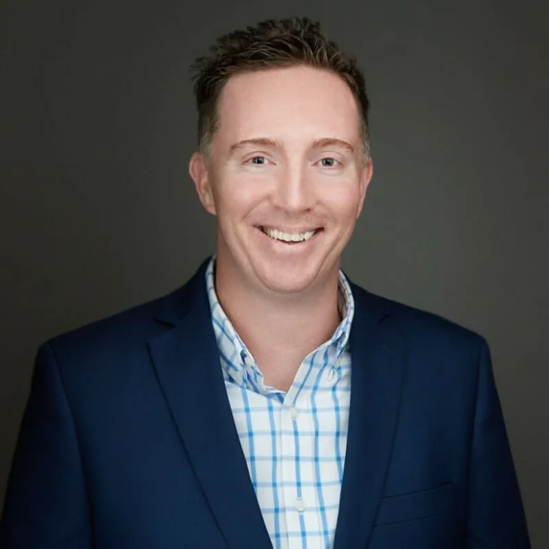 A man with short brown hair wearing a navy blue blazer and a blue-and-white checkered shirt smiles in front of a plain dark gray background.