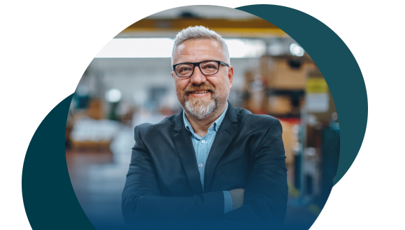 A smiling man with gray hair, beard, and glasses stands with arms crossed in a business suit, in an industrial or warehouse setting with blurred shelves and equipment in the background.