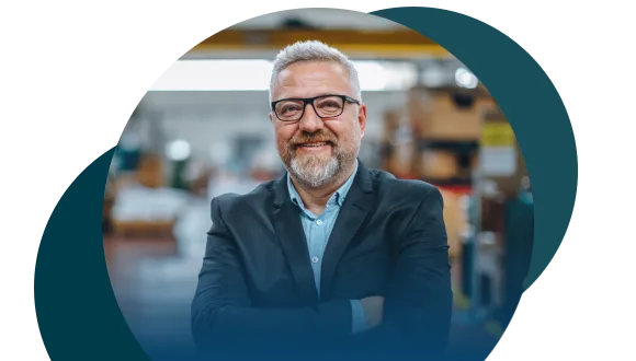 A smiling man with gray hair, beard, and glasses stands with arms crossed in a business suit, in an industrial or warehouse setting with blurred shelves and equipment in the background.