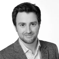 A man with short dark hair and a beard, wearing a light-colored shirt and a textured blazer, smiles slightly while facing the camera. The image is in black and white with a plain background, reflecting the professional spirit of Sustainability at Cority.