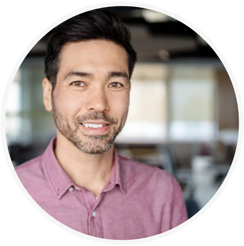 A man with short dark hair and a beard, wearing a pink button-up shirt, smiles at the camera. The background is a blurred office setting.